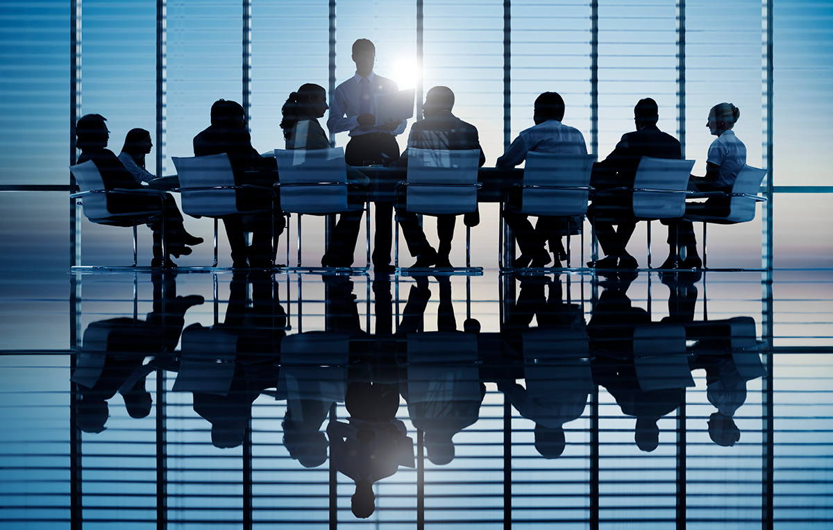 Board of directors meeting around a boardroom table, silhouetted against glass office windows