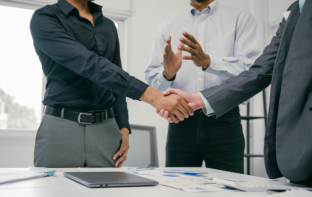 Two executives shake hands over documents at a conference table as a third looks on, closing a distressed mergers and acquisitions sale.